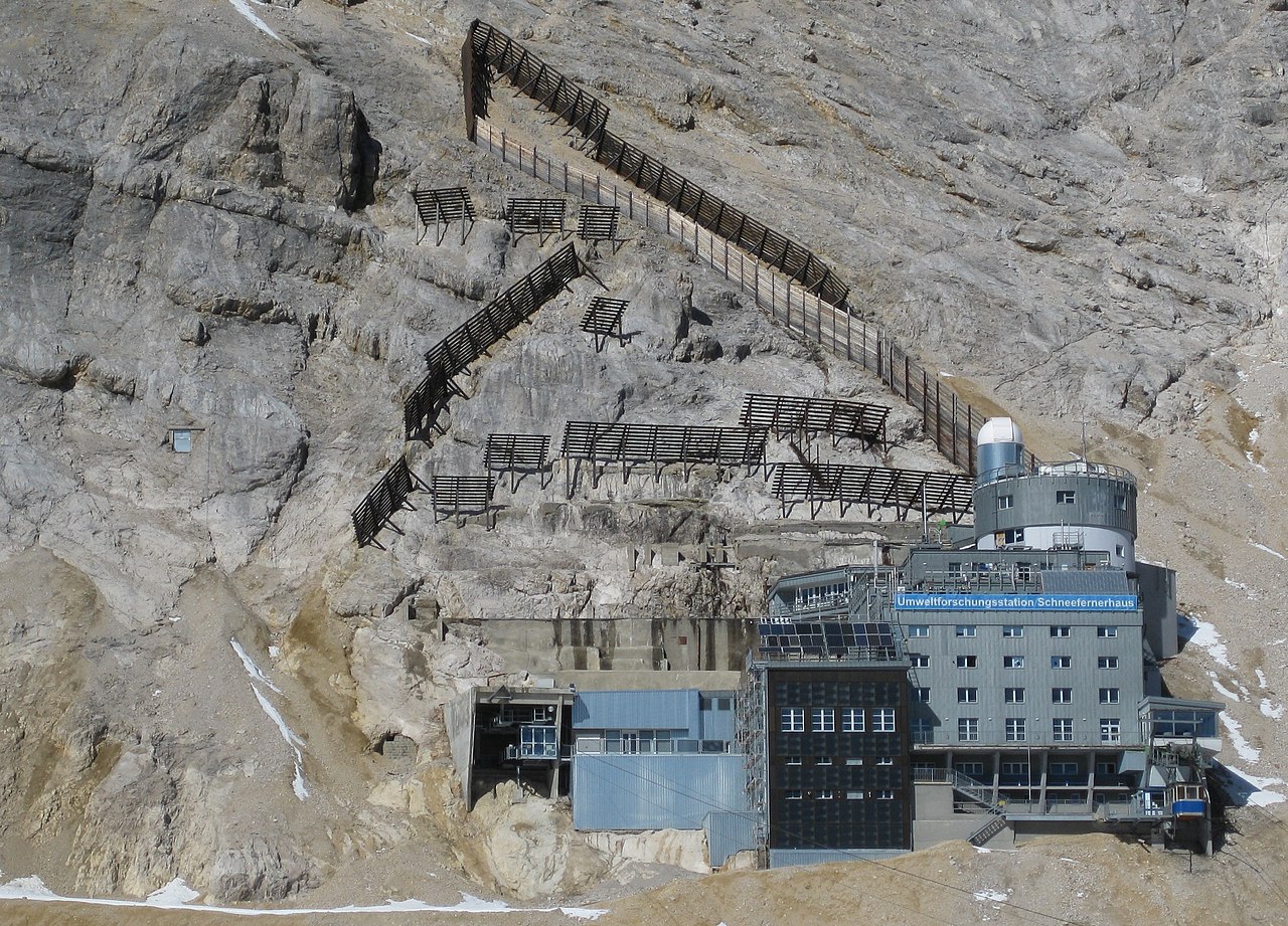 The Environmental Research Station Schneefernerhaus on the south side of the Zugspitze at 2,650 m. A multi-storey building with a white dome, a viewing platform on the roof, and snow fences on the slope above. The image is a 2010 photograph; the AllSkyCAM cluster that IFEX installed in 2025 is on a terrace not visible from this angle.