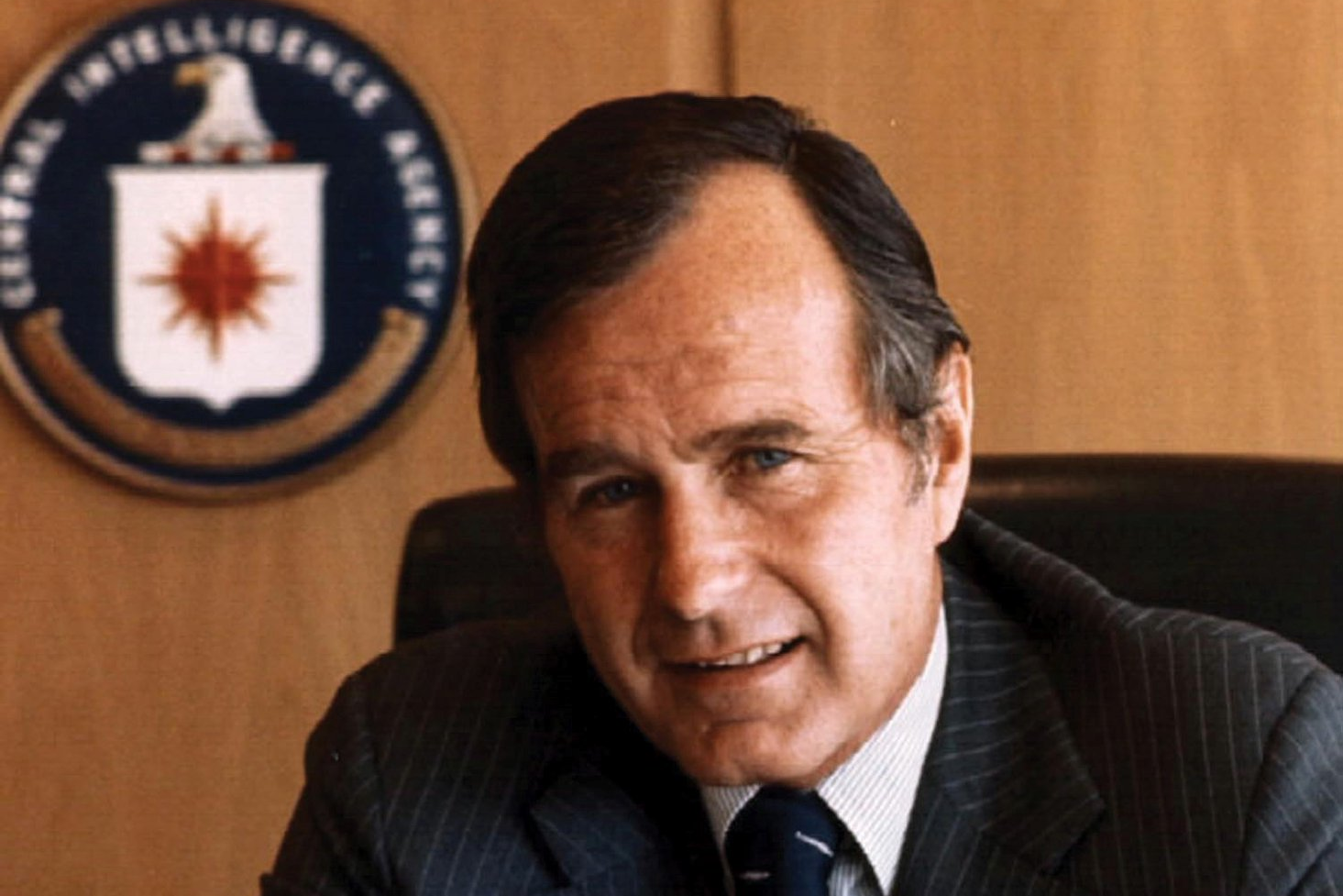 Color photograph of George H.W. Bush at his desk at CIA headquarters, with the agency seal visible behind him, taken during his tenure as Director of Central Intelligence from January 1976 to January 1977.