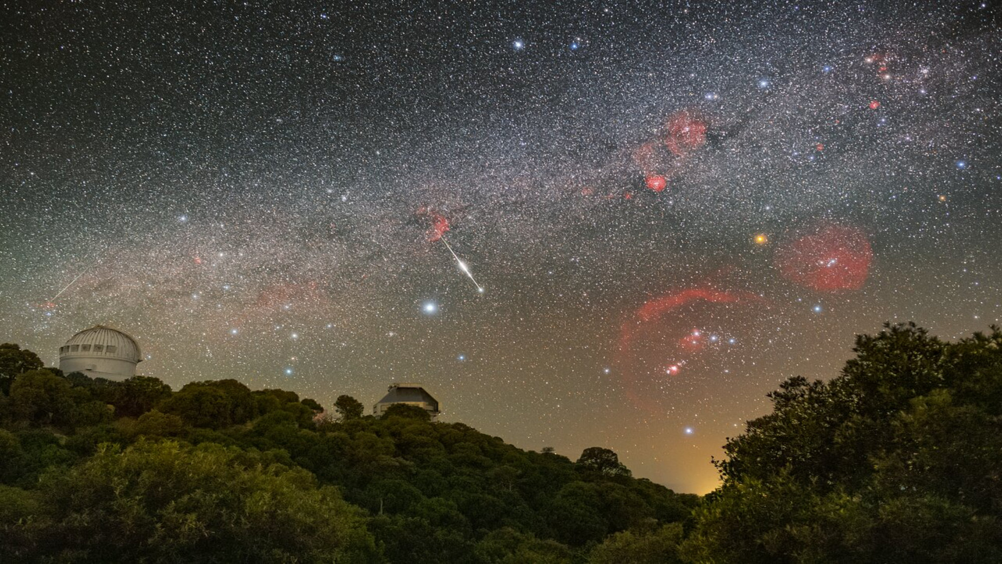 Wide-field astrophotograph of the Perseid meteor shower: multiple bright meteor streaks cross a star-filled Milky Way sky above two observatory domes and a forested ridge.