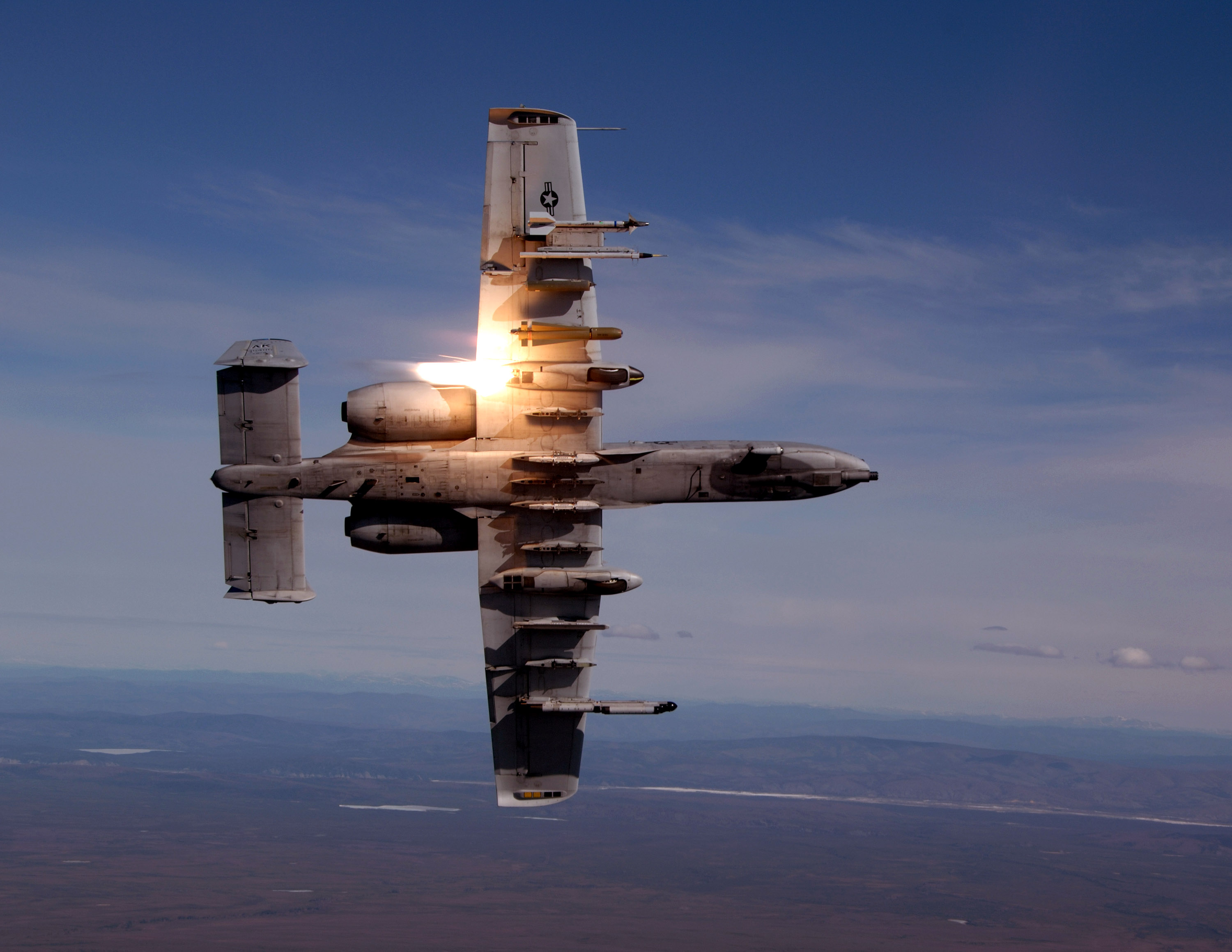 A single parachute-suspended illumination flare burning at sunset, falling away from the wing pylon of an A-10 Thunderbolt II in flight. The flare's magnesium-based light is a bright orange-yellow against a pale blue sky, with the dark silhouette of a mountain range below.
