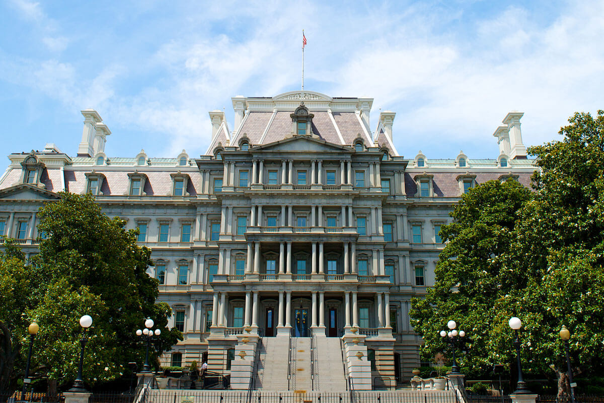 The Eisenhower Executive Office Building on the White House complex, photographed from Pennsylvania Avenue with mature trees framing the central bay, the U.S. flag above the main entrance, and the building's full granite facade visible.
