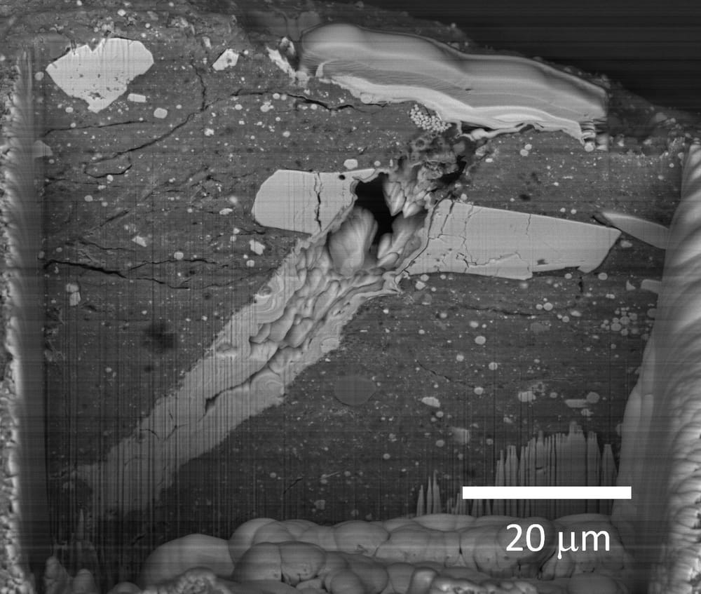 Scanning electron microscope image of a micrometeorite impact crater in a particle of asteroid Bennu material, with a 20-micrometre scale bar.