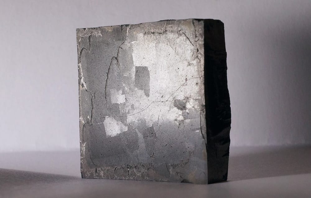 A small dark ceramic YBCO (yttrium barium copper oxide) high-temperature superconductor specimen on a neutral grey surface, photographed in a clean documentary style. Real specimen, not Ning Li's hardware.