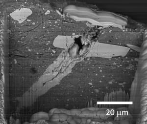 Scanning electron microscope image of a micrometeorite impact crater in a particle of asteroid Bennu material, with a 20-micrometre scale bar.