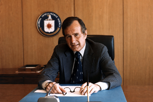 George H.W. Bush at his CIA headquarters desk during his 1976–77 tenure as Director of Central Intelligence, with the CIA seal behind him. The public-domain archival photograph is presented in a landscape layout with only the side background extended.
