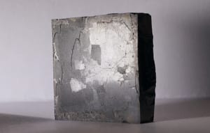 A small dark ceramic YBCO (yttrium barium copper oxide) high-temperature superconductor specimen on a neutral grey surface, photographed in a clean documentary style. Real specimen, not Ning Li's hardware.