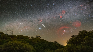 Wide-field astrophotograph of the Perseid meteor shower: multiple bright meteor streaks cross a star-filled Milky Way sky above two observatory domes and a forested ridge.