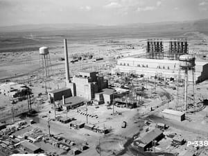 The B Reactor at the Hanford Site, Washington State, photographed from the southeast showing the 28-by-46-metre reactor building, the 38-metre ventilation stack, the 185-B deaerating plant towers, the water-treatment complex, and the Columbia River plateau in the background. The B Reactor was the first full-scale nuclear reactor ever built (startup 26 September 1944) and supplied the plutonium for the Trinity test and the Nagasaki bomb. It remained operational until 1968 and is a National Historic Landmark. Hanford is the western anchor of the U.S. atomic warfare complex used in the SCU 1945–1975 study.
